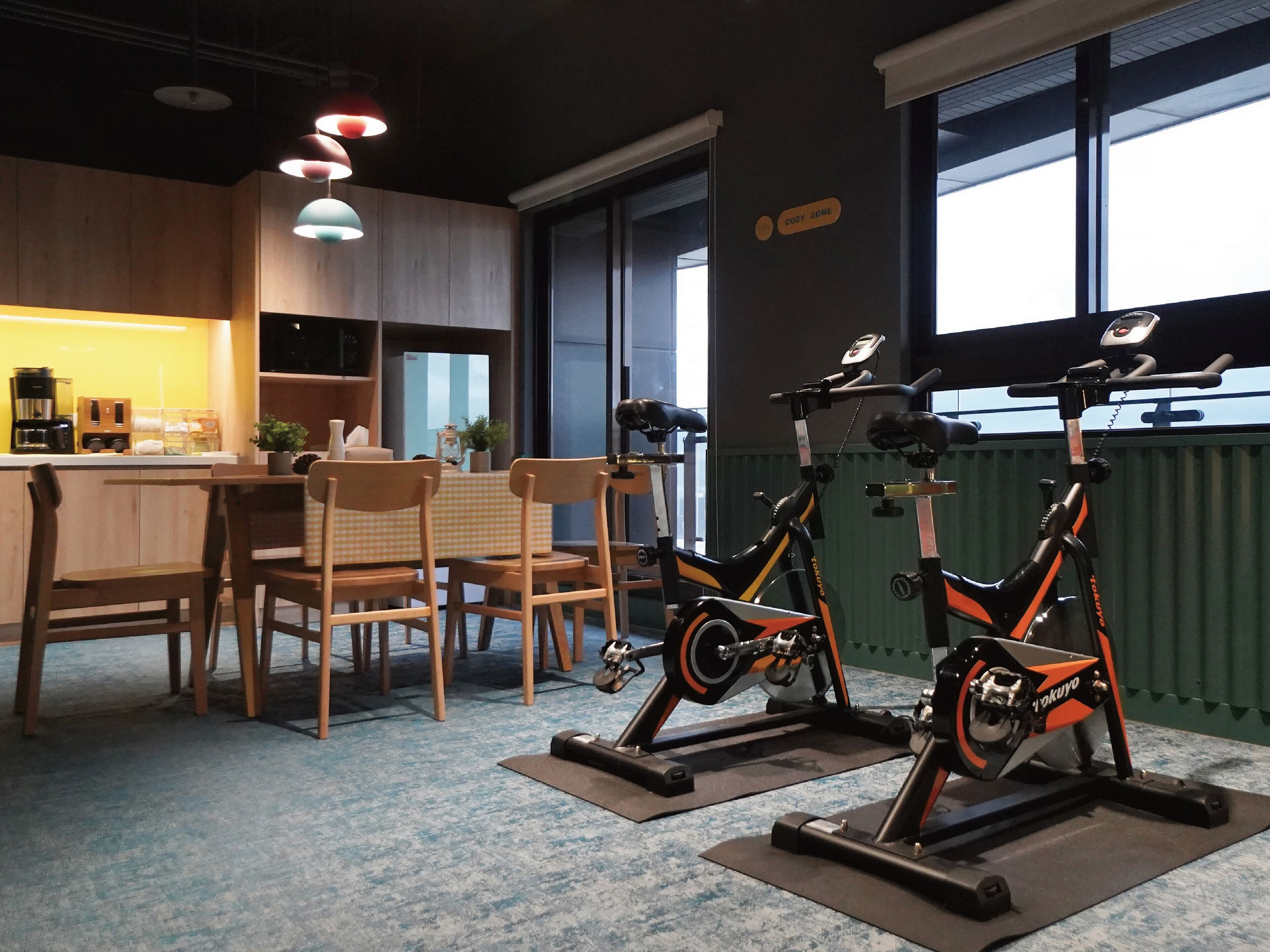 A room with a dining area, two exercise bikes, a kitchen, and a sign indicating a 'COZY ZONE'.