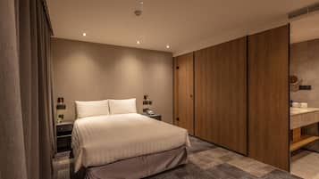 A modern hotel room with a large bed, a wooden wardrobe, a bedside table with a lamp, and a bathroom with a mirror and sink.