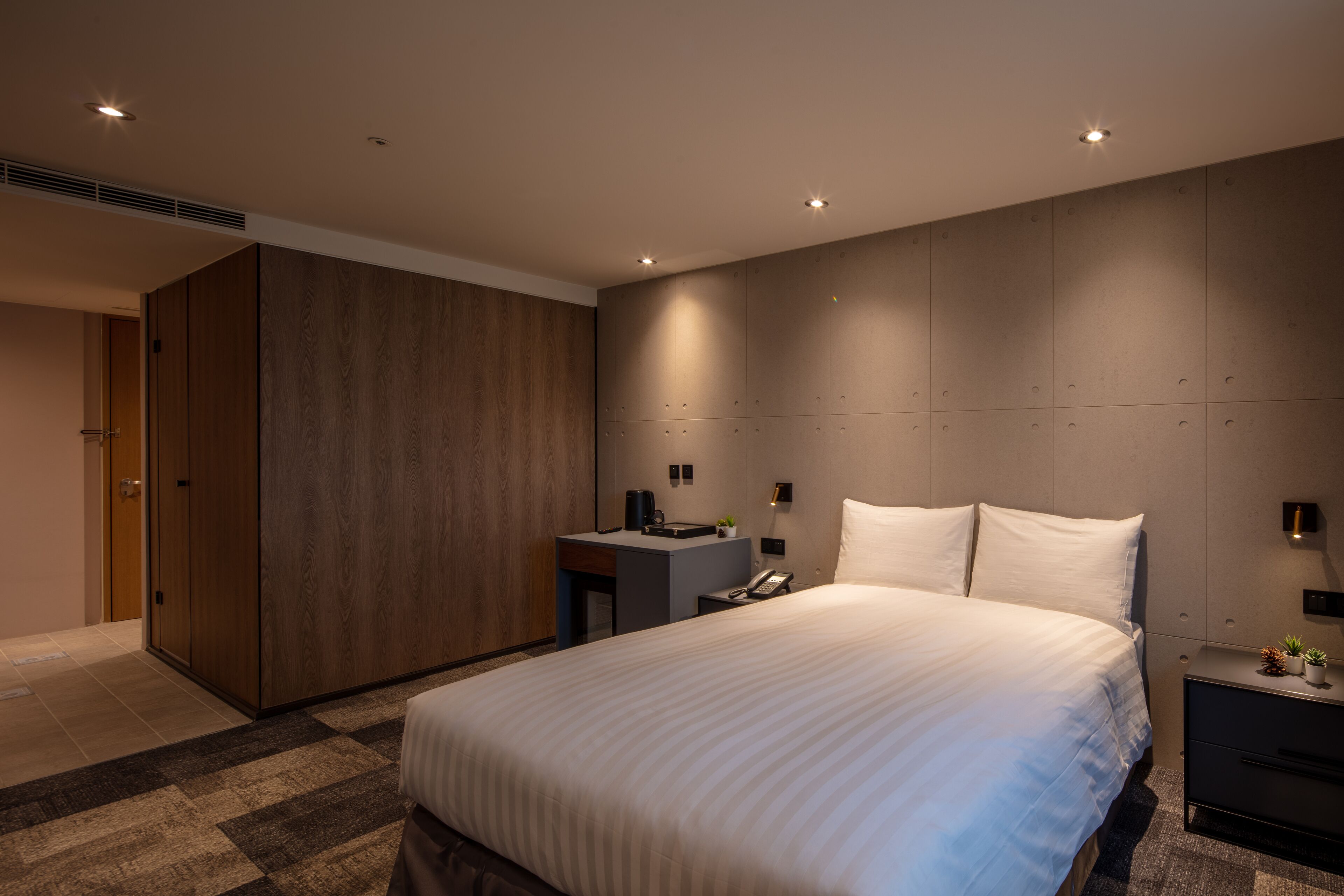 A modern hotel room with a large bed, a desk, a wardrobe, and a nightstand with a lamp.