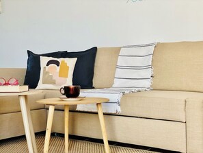Un sofá color beige con un cojín azul oscuro, un cojín blanco con un diseño gráfico y una manta a rayas. Frente al sofá, una pequeña mesa de madera con una taza y un libro.