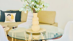 Une table à manger avec un plateau en verre, accompagnée de deux chaises blanches, d’un vase beige orné de feuillage vert et d’un canapé avec des coussins en arrière-plan.