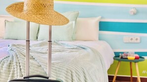 Un lit avec une tête de lit à rayures bleues et blanches, un chapeau en osier posé sur un présentoir et une valise bleue sur le sol.