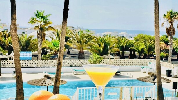 Une scène au bord de la piscine, avec une vue dégagée sur la mer, un verre à martini contenant une boisson jaune et deux oranges posées sur un journal.