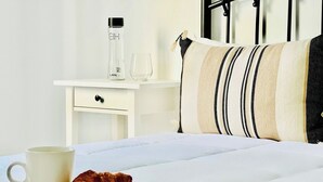 Una cama bien hecha con un almohadón, una mesita de noche con una botella y un vaso, y un croissant sobre un posavasos de madera.