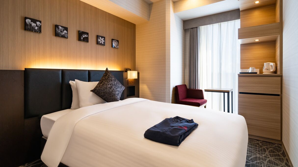 Quarto de hotel com cama, uma cadeira vermelha, uma mesinha, máquina de café e prateleira na parede.