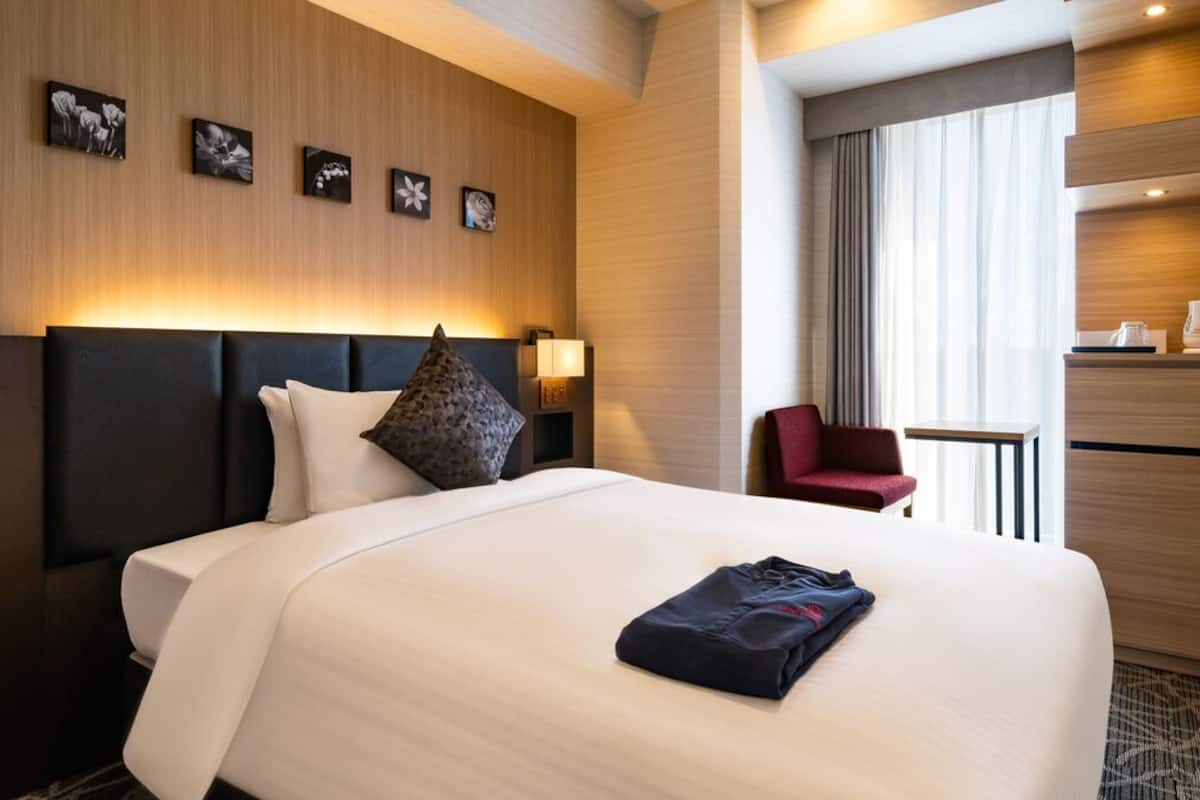 A hotel room with a bed, a red chair, a small table, a coffee maker, and a wall-mounted shelf.