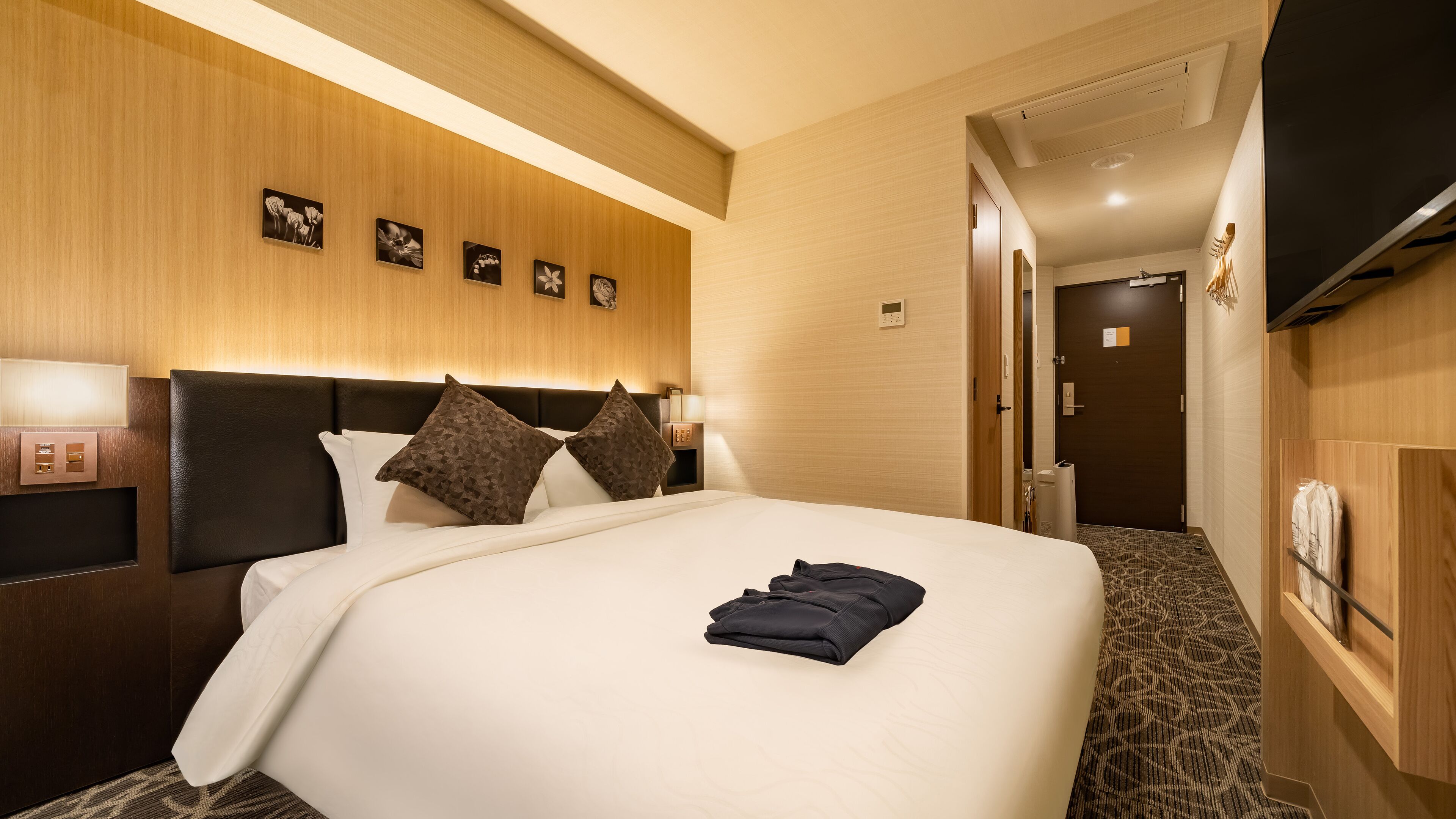 Habitación de hotel con una cama grande, cabecera, mesita de noche con lámpara, panel de control en la pared y puerta que da al baño.