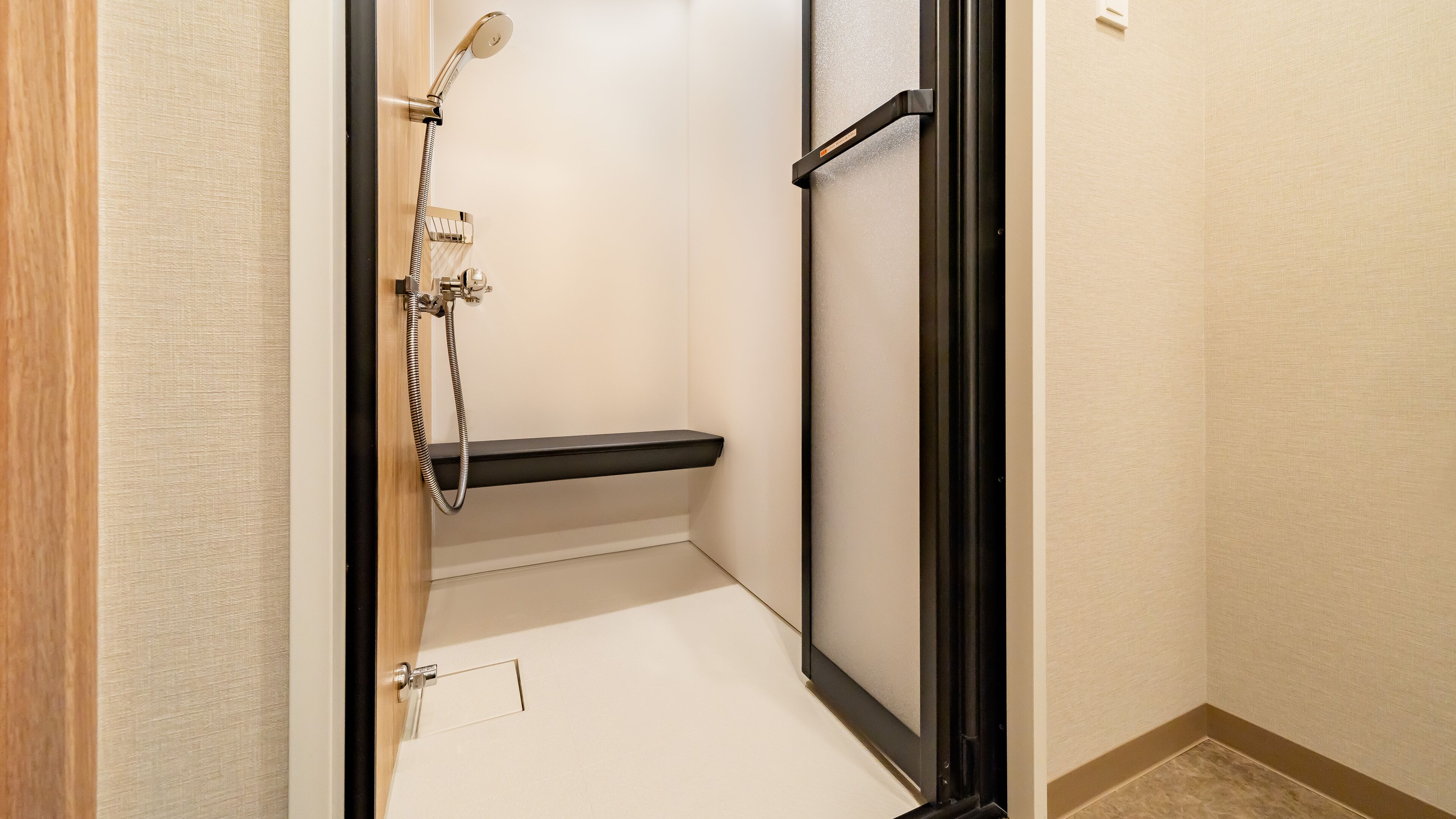 A small, enclosed shower area with a showerhead and hose, a black shelf, and a door with a handle.