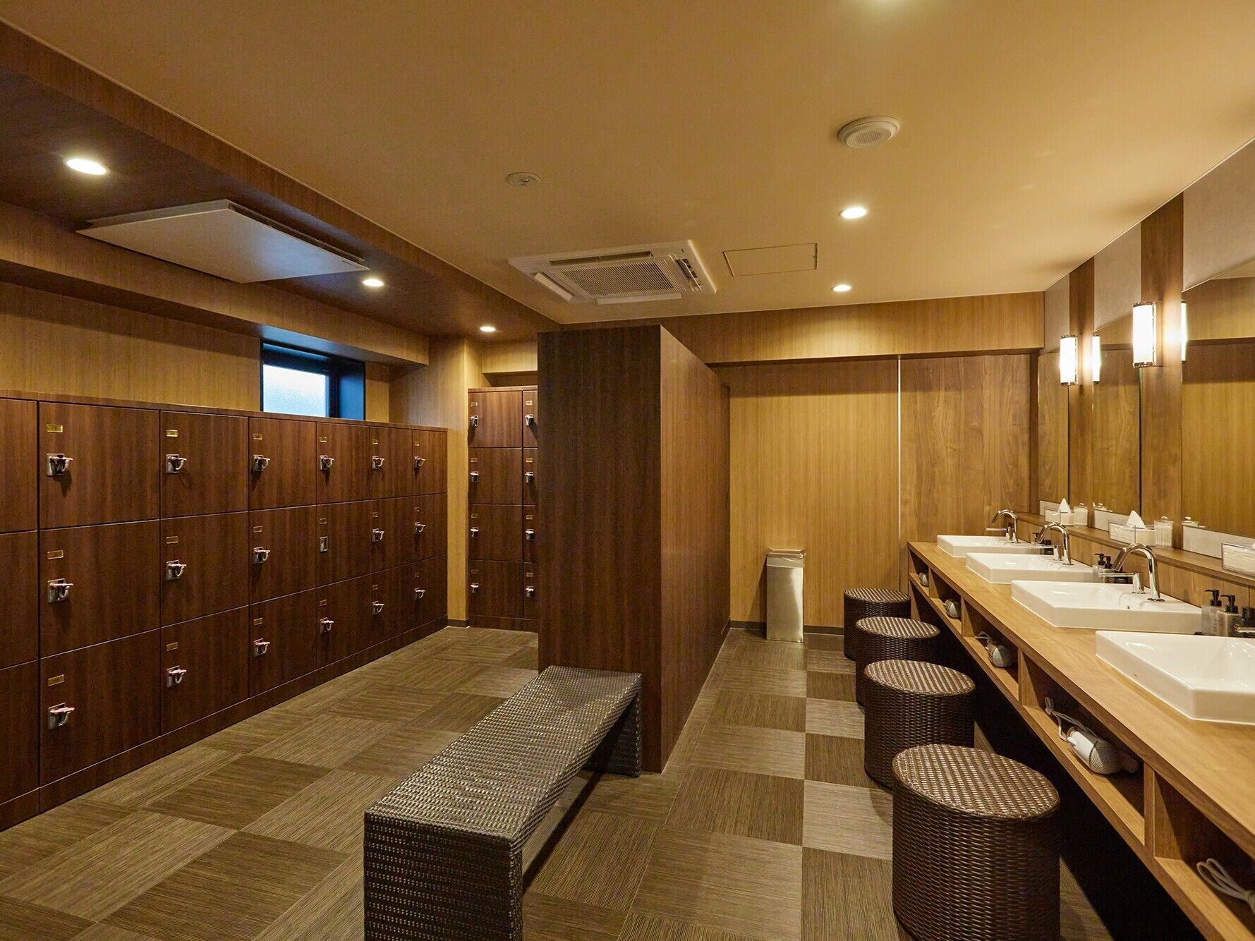 A hotel bathroom with multiple lockers, a bench, and a row of sinks with mirrors.