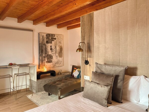 Un dormitorio moderno con techo de madera, un cuadro abstracto grande, una cama con muchas almohadas, una mesita con sillas y una lámpara de pared.