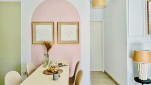 A dining area with a long table, chairs, and a vase with dried flowers. There are framed artworks on the wall and a lamp on a side table.