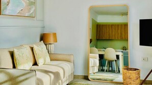 A living room with a sofa, a lamp, a large mirror reflecting a dining area with a table and chairs, and a kitchen visible through the mirror.