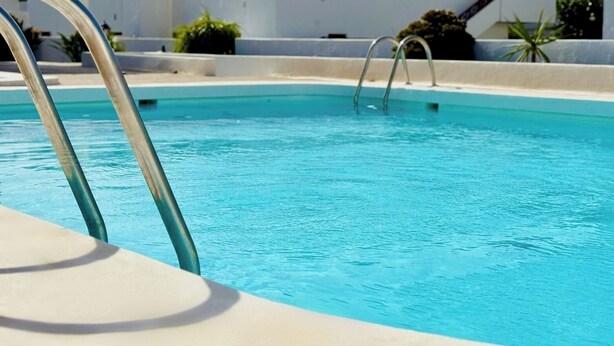 A clear blue swimming pool with metal ladders, surrounded by a white pool deck and some greenery. - Canaryislandshost l Sol (Puerto del Carmen)
