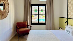 A hotel room with a bed, a chair, a window with a view of a pool, and a hanging light fixture. - Canaryislandshost l Sol (Puerto del Carmen)
