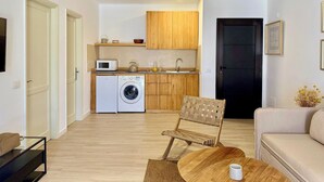 A compact living space with a kitchenette, a washing machine, a wooden chair, a sofa, and a coffee table. - Canaryislandshost l Sol (Puerto del Carmen)