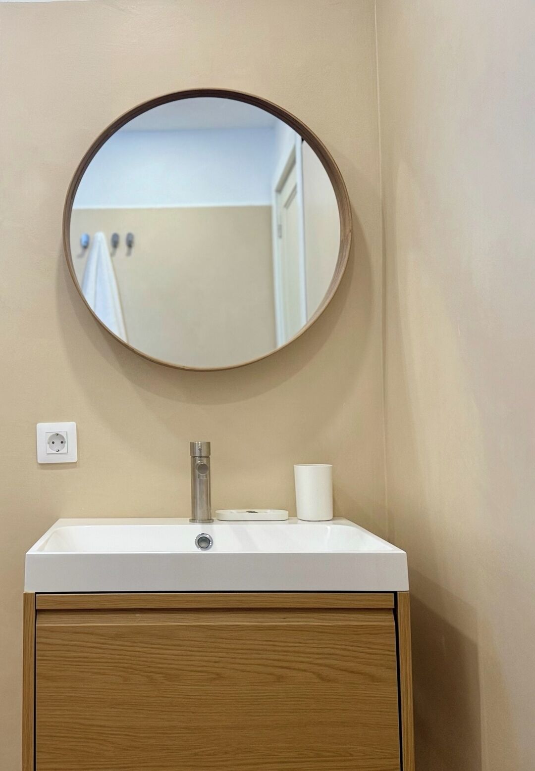 A bathroom with a round mirror, a white sink, a faucet, a toilet paper holder, and a wooden cabinet.