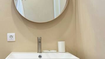 A bathroom with a round mirror, a white sink, a faucet, a toilet paper holder, and a wooden cabinet.