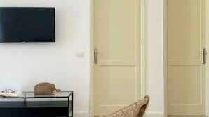 A room with two closed doors, a mounted television, a small table with a pair of glasses and a cap, and a chair. - Canaryislandshost l Sol (Puerto del Carmen)