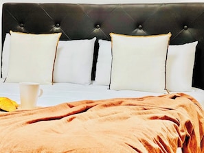 Un lit avec une tête de lit capitonnée, des oreillers blancs, une couverture marron et une tasse blanche.