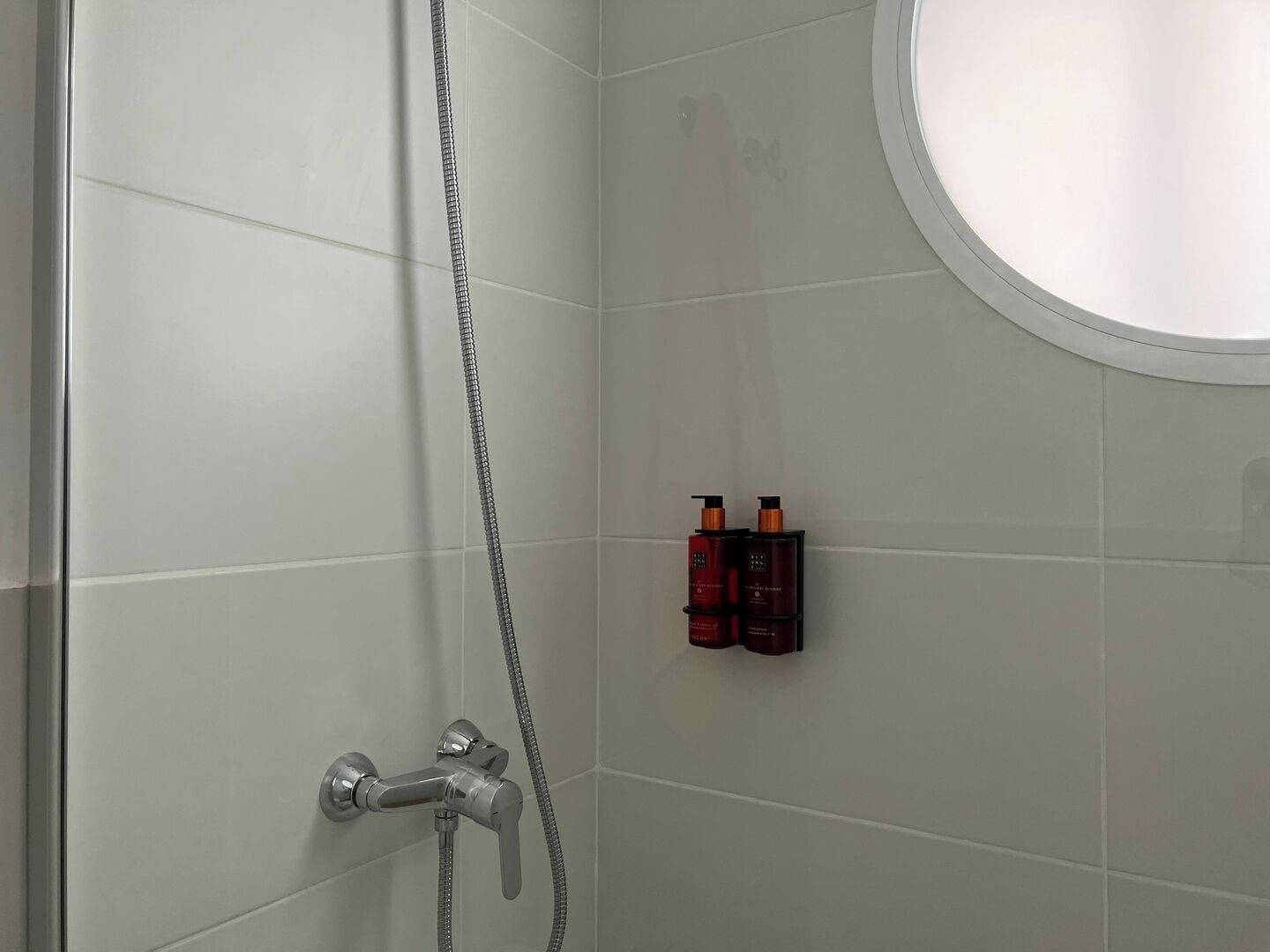 Une douche équipée d’un pommeau de douche à main et d’un robinet mural. Deux flacons de shampoing et d’après-shampoing sont posés sur une petite étagère.
