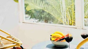 A table with a bowl of fruit and two glasses of red wine, a wicker chair, and a window with palm trees outside. - Canaryislandshost l Mar Lanzarote (Puerto del Carmen)