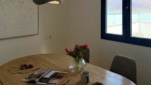 Un coin repas avec une table en bois, un vase rempli de fleurs rouges, une tasse et une vue sur une étendue d’eau par la fenêtre.