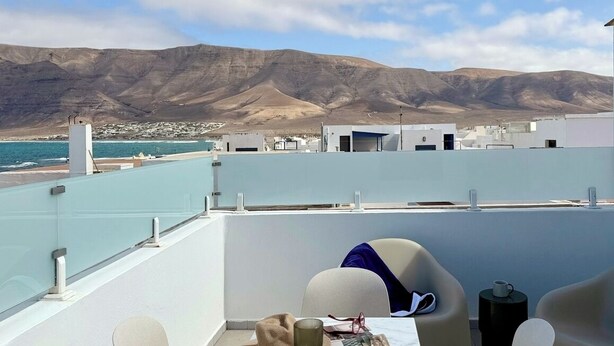 Une terrasse sur le toit, avec une table dressée pour deux personnes, un chapeau et un verre à vin. La terrasse offre une vue sur une ville côtière et sur les montagnes.