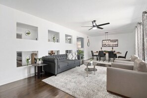 En moderne stue med en grå sofa, et rundt sofabord og et spiseområde med en lysekrone.
