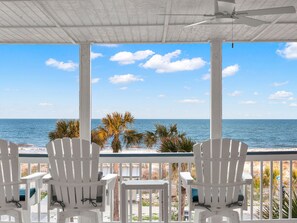 A covered outdoor seating area with white chairs and a ceiling fan, overlooking a beach with palm trees and the ocean. - Gorgeous Ocean Views from Balcony (Tybee Island)