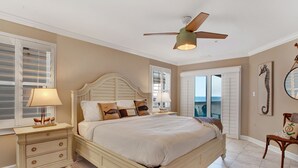 Een slaapkamer met een bed, nachtkastjes, een plafondventilator, een raam met jaloezieën, een deur naar een balkon en een kunstwerk van een zeepaardje aan de muur.