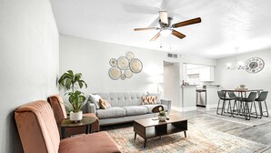 Una sala de estar moderna con un sofá gris, un sillón marrón, una mesa de centro y una zona de comedor con una lámpara de araña y un reloj en la pared.