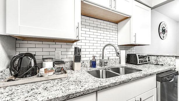 Una cocina moderna con armarios blancos, encimera de granito, pileta de acero inoxidable, un tostador y un reloj en la pared.