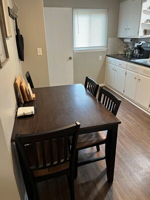 Un coin repas avec une table et des chaises en bois, une fenêtre avec des stores et une cuisine dotée de meubles blancs et d’un plan de travail noir.