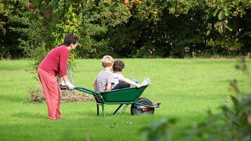 A person in a red shirt and pink pants pushing a green wheelbarrow with two children sitting inside.