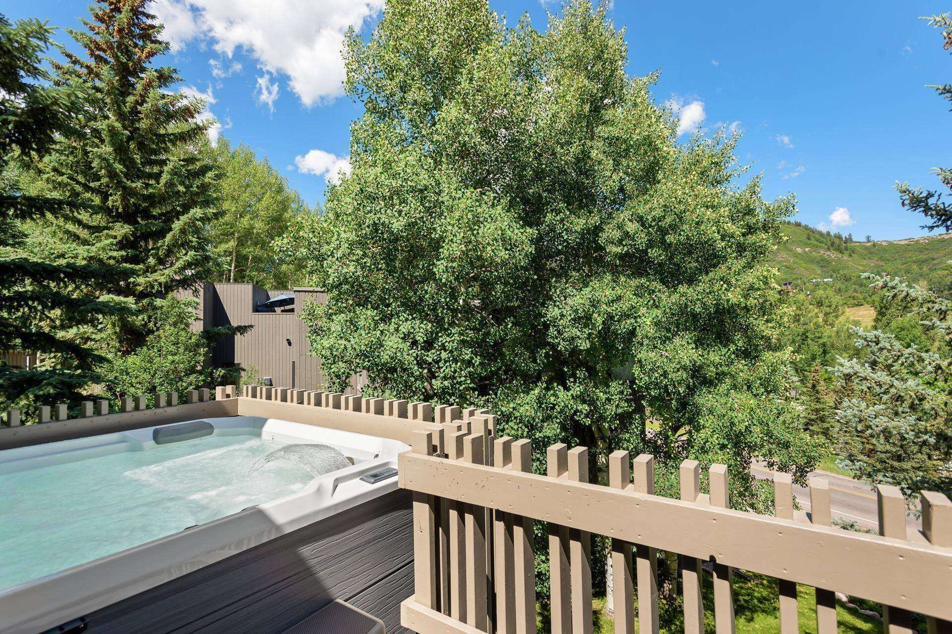 A hot tub on a wooden deck with a wooden fence, surrounded by trees and a building in the background.