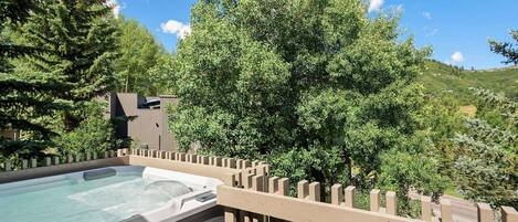 A hot tub on a wooden deck with a wooden fence, surrounded by trees and a building in the background.