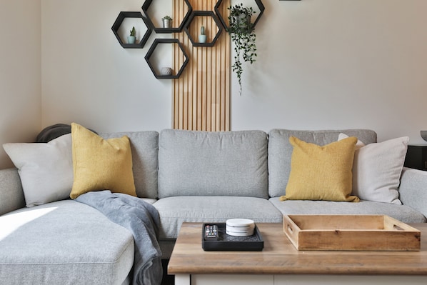 Ein modernes Wohnzimmer mit einem grauen Ecksofa, gelben Kissen, einem Holztisch und sechseckigen Wandregalen mit Pflanzen.