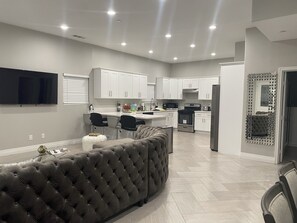 Una cocina moderna con armarios blancos, un refrigerador de acero inoxidable y un horno empotrado. Hay un sofá tapizado en gris y una zona de comedor con una mesa y sillas blancas.