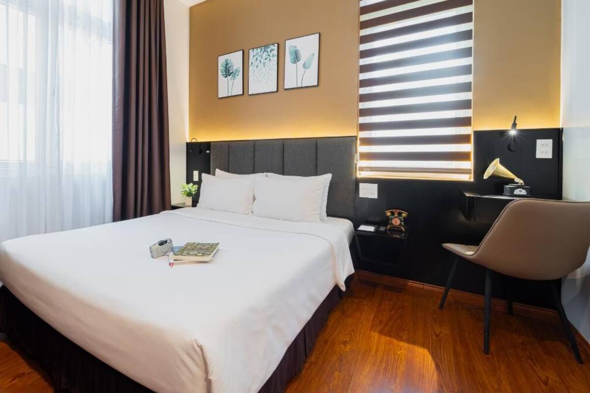A hotel room with a large bed, a desk with a lamp, and a window with blinds.