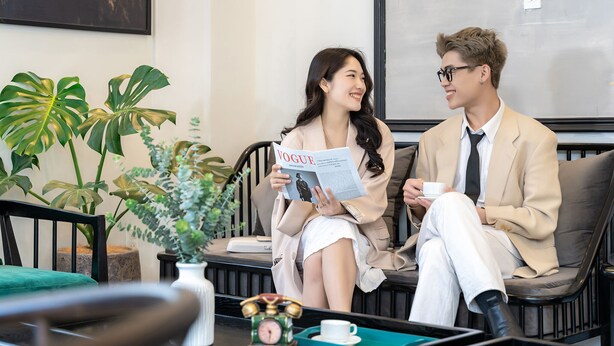 Deux personnes sont assises sur un canapé, l’une tient un magazine, et une horloge ancienne est posée sur une table.