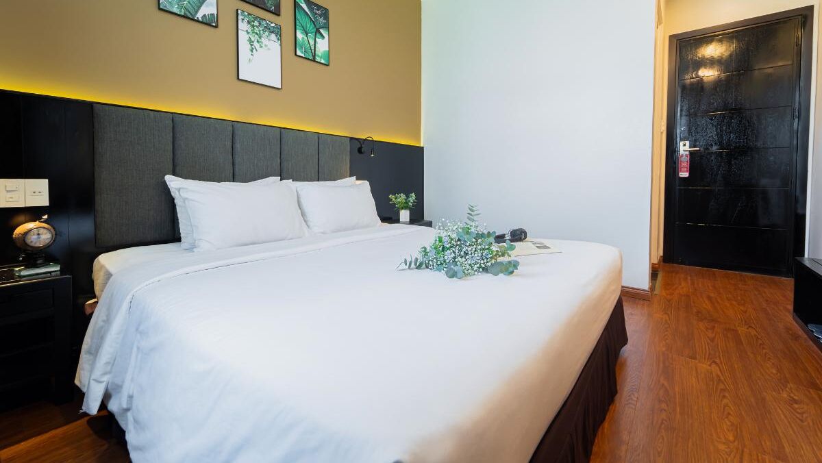 A neatly made bed with white linens, a bedside table with a clock, and a wall-mounted light.