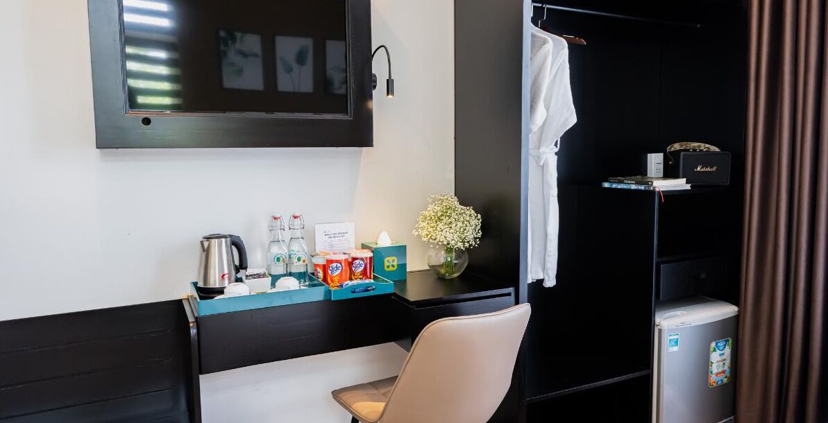 A hotel room with a bed, a desk with a chair, a small fridge, a television mounted on the wall, and a window with curtains.
