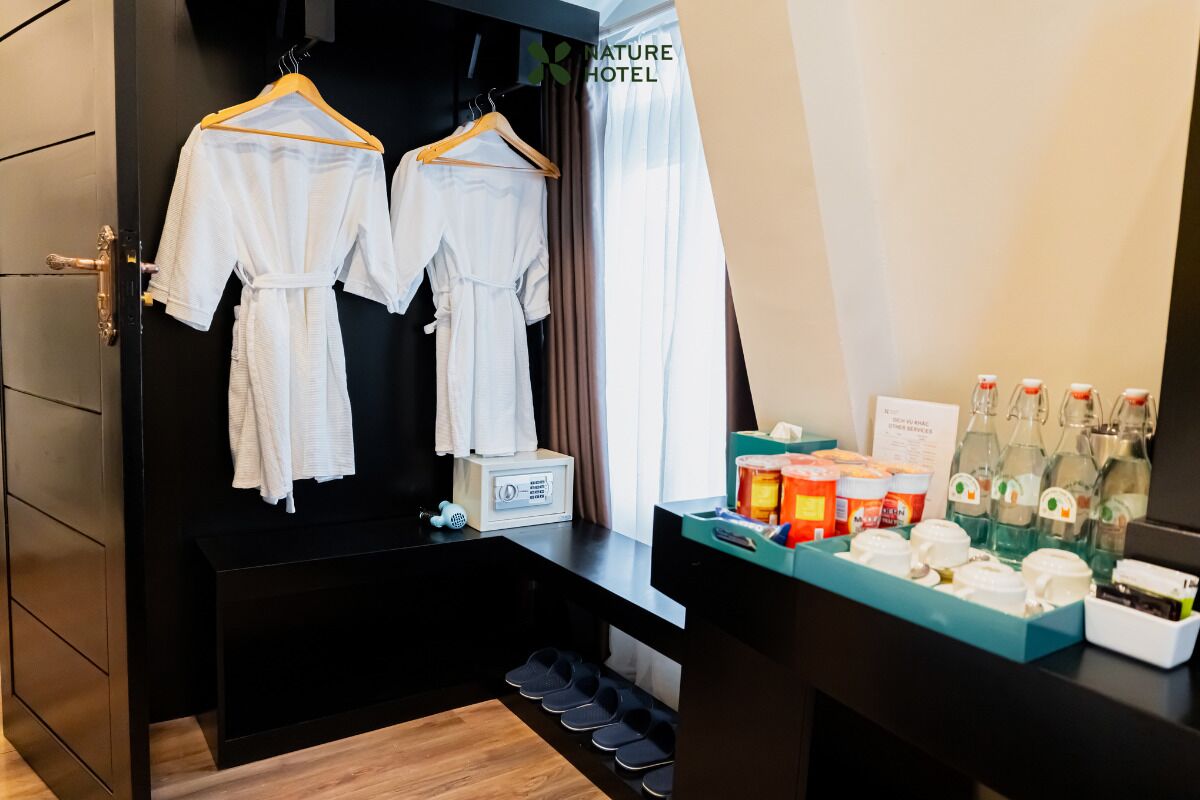 A hotel room with a black wardrobe, two white robes hanging on a wooden hanger, a vanity with toiletries, and a window with curtains.