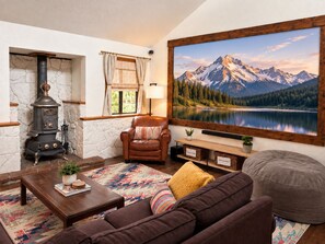 A cozy living room with a stone wall, a wood-burning stove, a leather armchair, a sofa, a coffee table, and a large mountain landscape painting.