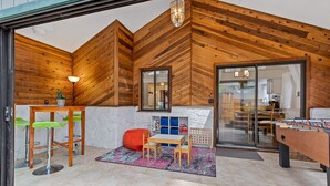 A cozy room with wooden walls, a foosball table, a small table with chairs, and a glass door leading to another room.