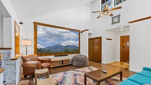A spacious living room with a large window, leather chairs, a blue sofa, a wooden coffee table, and a view of mountains.