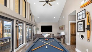A pool table in a spacious room with a ceiling fan, a mounted TV, and a view of the outdoors through large windows. - AMAZING Lakeview, Private Putting Green, Hot Tub, Pools, Pool Table, Pickleball (Branson)