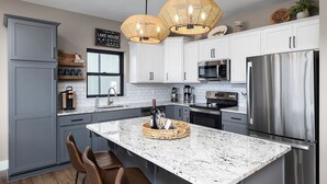 A modern kitchen with white cabinets, stainless steel appliances, a granite island, and wicker pendant lights. - AMAZING Lakeview, Private Putting Green, Hot Tub, Pools, Pool Table, Pickleball (Branson)