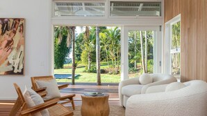 A spacious living room with a large sliding glass door leading to a lush garden. There are two wooden chairs and a round wooden coffee table. - Miren Estate - lavish hinterland estate with pool (Federal)