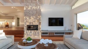 A modern living room with a stone fireplace, a flat-screen TV, and a dining area in the background. - Miren Estate - lavish hinterland estate with pool (Federal)